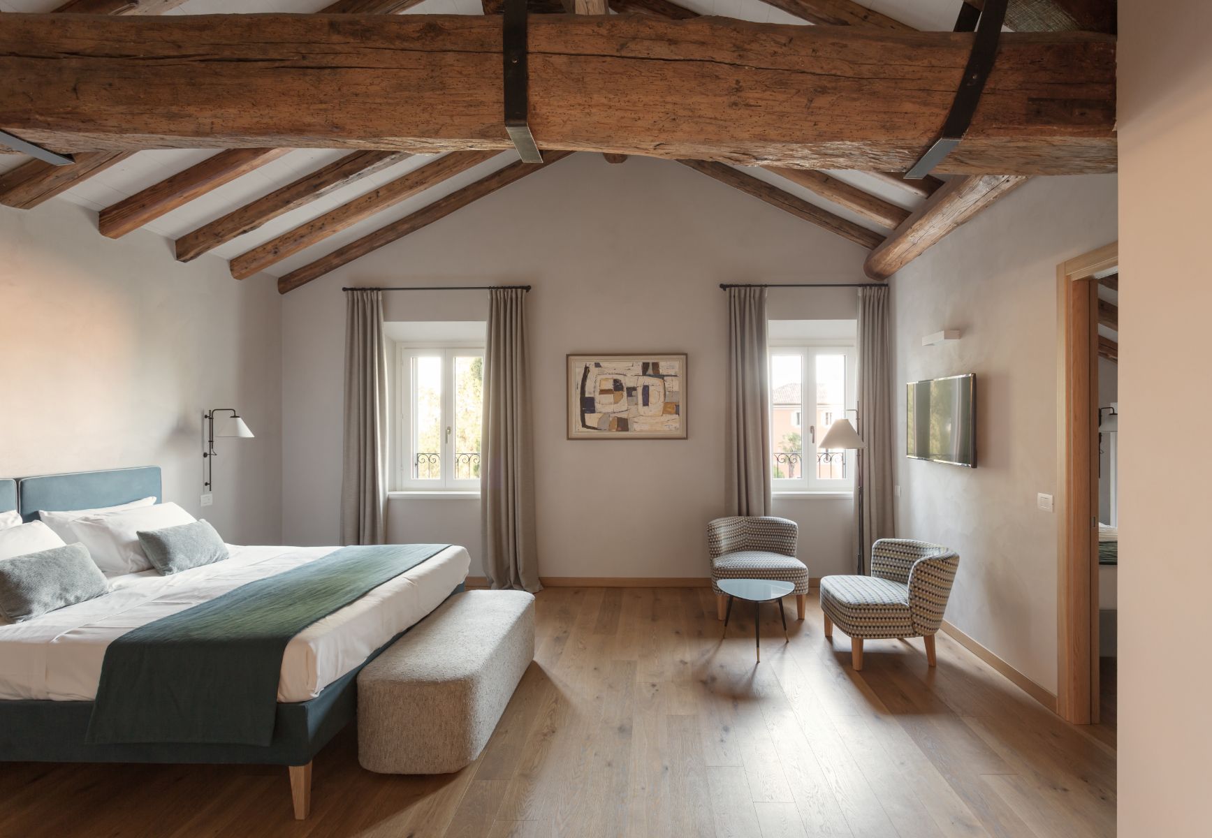Roncolo 1888, Europe — Spacious bedroom with exposed wooden beams, blue upholstered bed, woven armchairs, dual windows with linen curtains, light wood flooring, minimalist decor, and soft natural light.