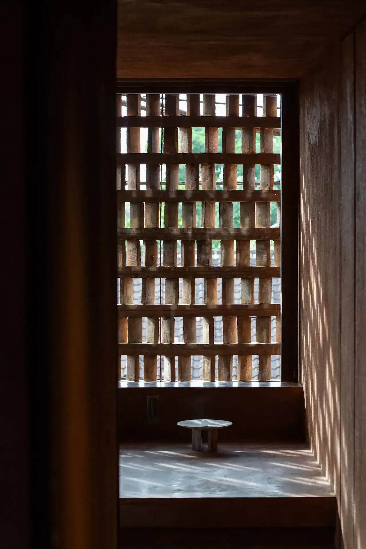 Further Hotel, Asia — Wooden lattice window framing garden views, natural light casting geometric shadows across concrete floor and minimalist stone table.