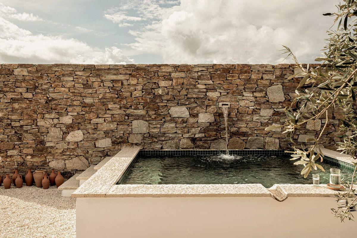The Rooster, Europe — Sunlit stone-walled pool courtyard with olive tree, terracotta vessels, and minimalist architectural design.