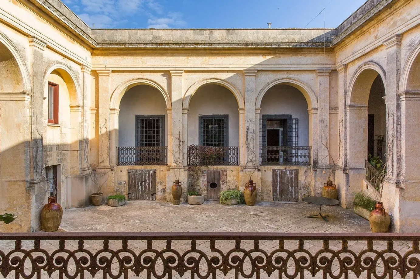 Palazzo Daniele, Europe — Sunlit courtyard with arched colonnades, weathered limestone facades, terracotta vessels, and wrought-iron balconies.