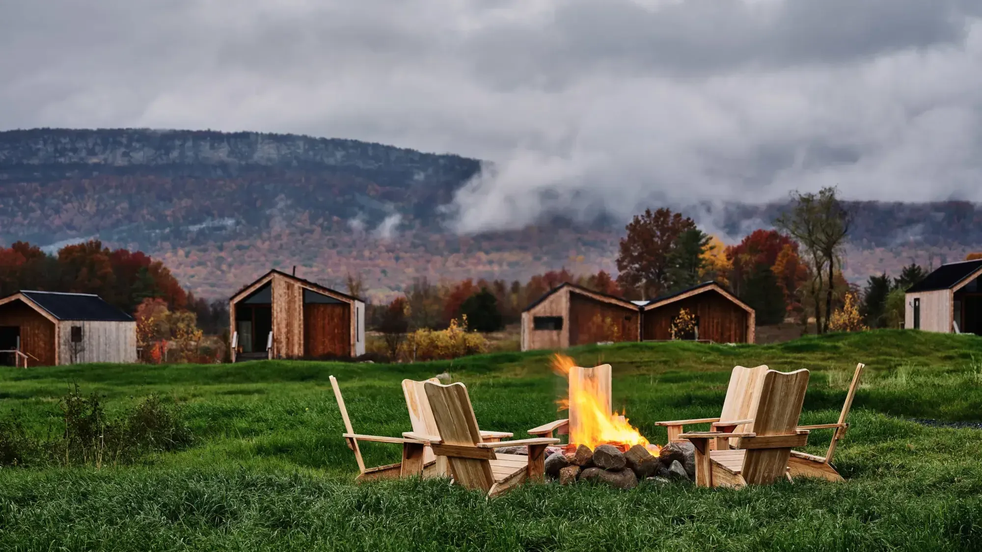 Wildflower Farms, North America — Adirondack chairs beside a crackling fire pit overlooking autumn-colored valleys and weathered farm buildings at dusk.