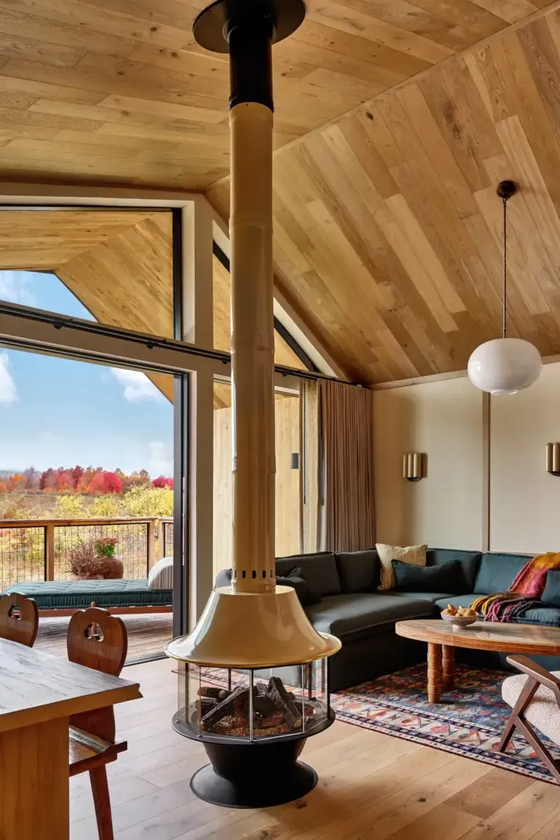Wildflower Farms, North America — Modern wood-lined living room with suspended fireplace, black sectional sofa, and floor-to-ceiling windows overlooking autumn foliage.