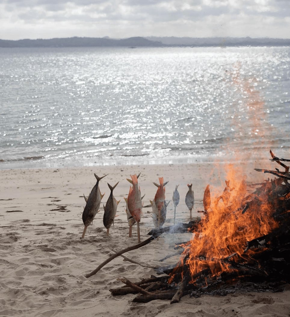 Innit Lombok, Indonesia — Fish roasting over driftwood fire on beach at sunset, with shimmer of sea and forested coastline beyond.