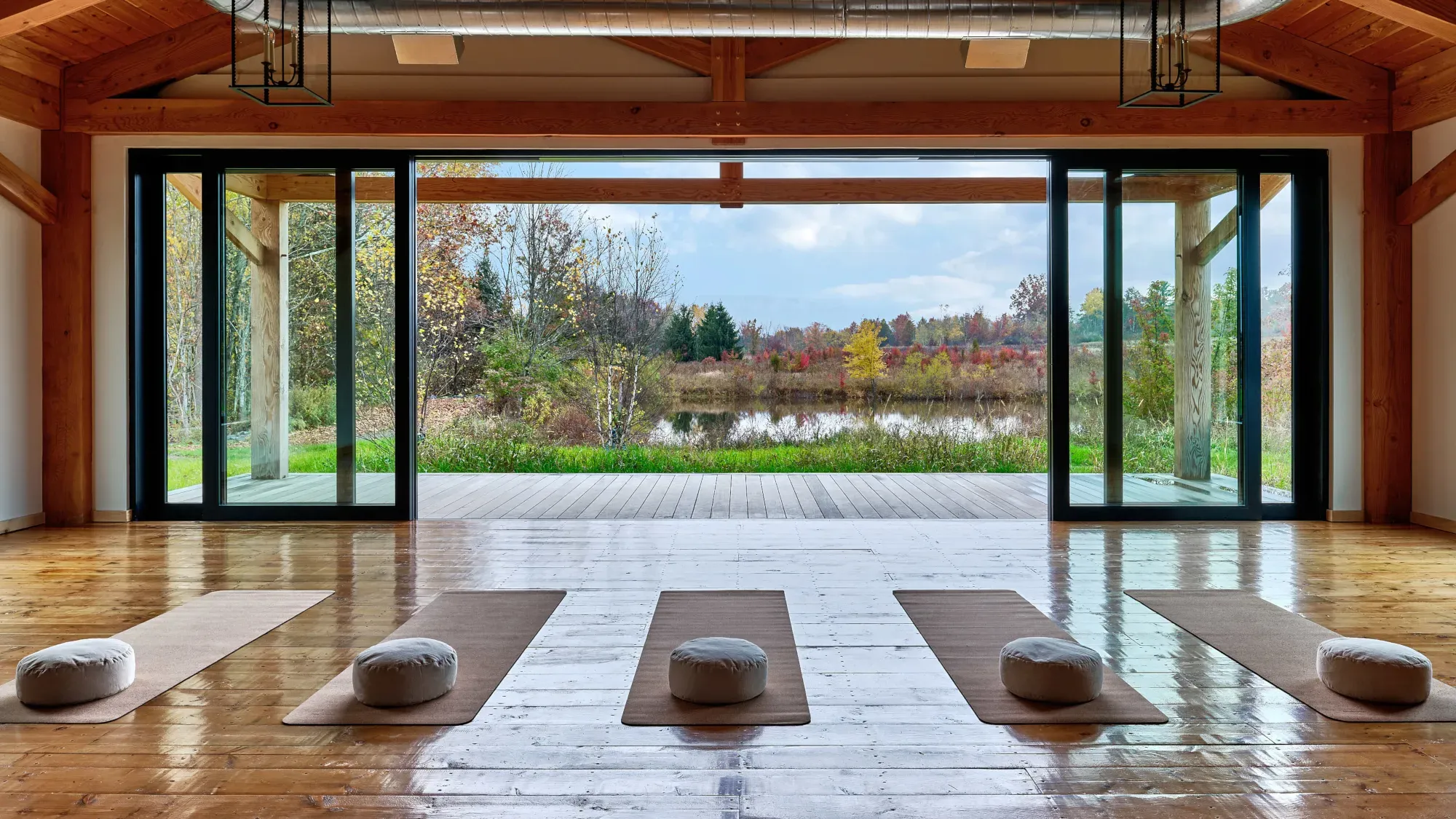 Wildflower Farms, North America — Minimalist yoga studio with floor-to-ceiling windows overlooking wetlands, timber beams, meditation cushions on natural fiber mats, autumn landscape.