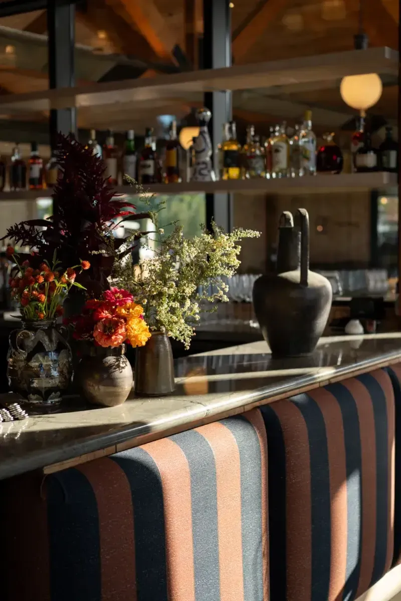 Wildflower Farms, North America — Luxury bar with striped upholstery, fresh floral arrangements, backlit liquor shelves, and warm ambient lighting.