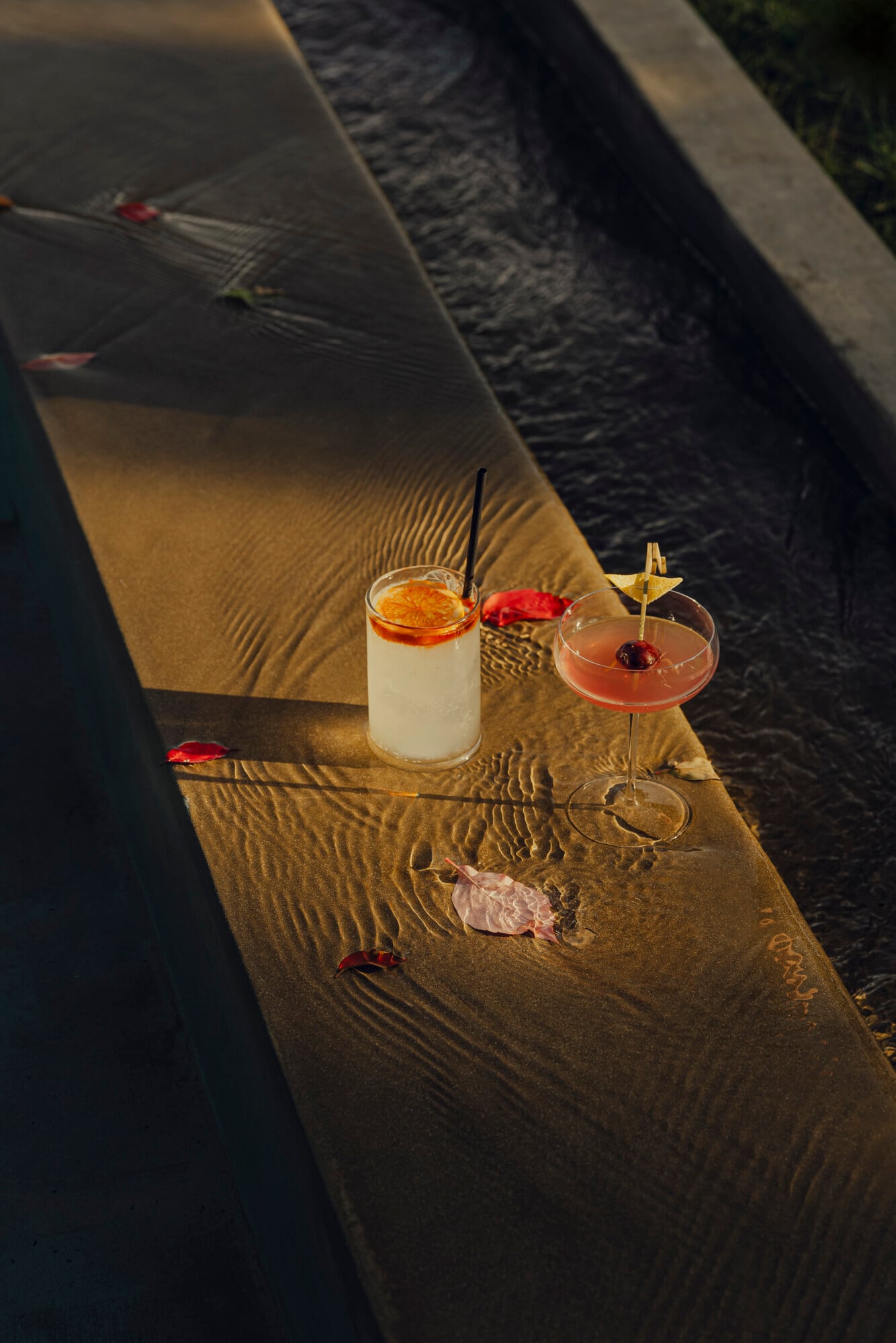 Pool Bar, Italy — Two cocktails with red garnishes and a tall glass drink on sand beside water, dappled sunlight casting ripple shadows, minimalist luxury hospitality setting.