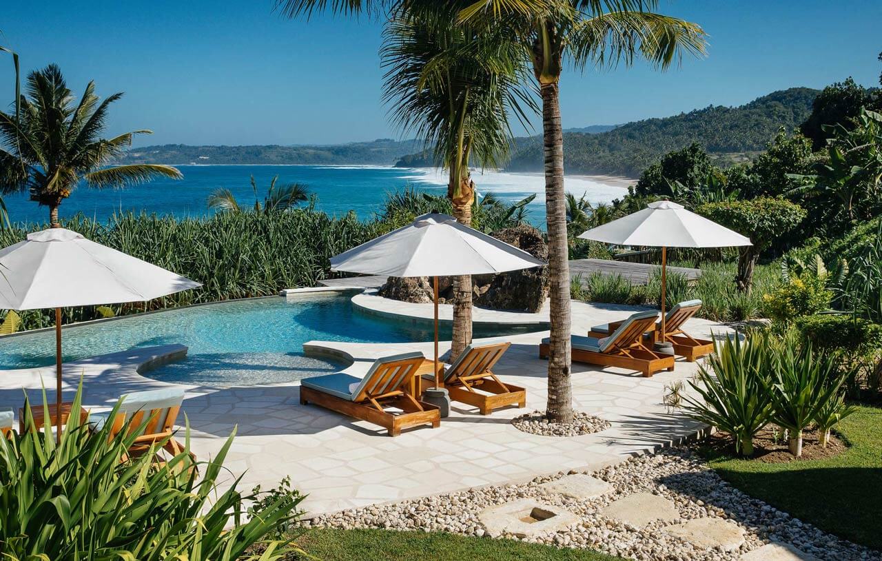 NIHI Sumba, Asia — Clifftop infinity pool with teak loungers, white umbrellas, and turquoise ocean vista overlooking white-sand beach and forested coastline.