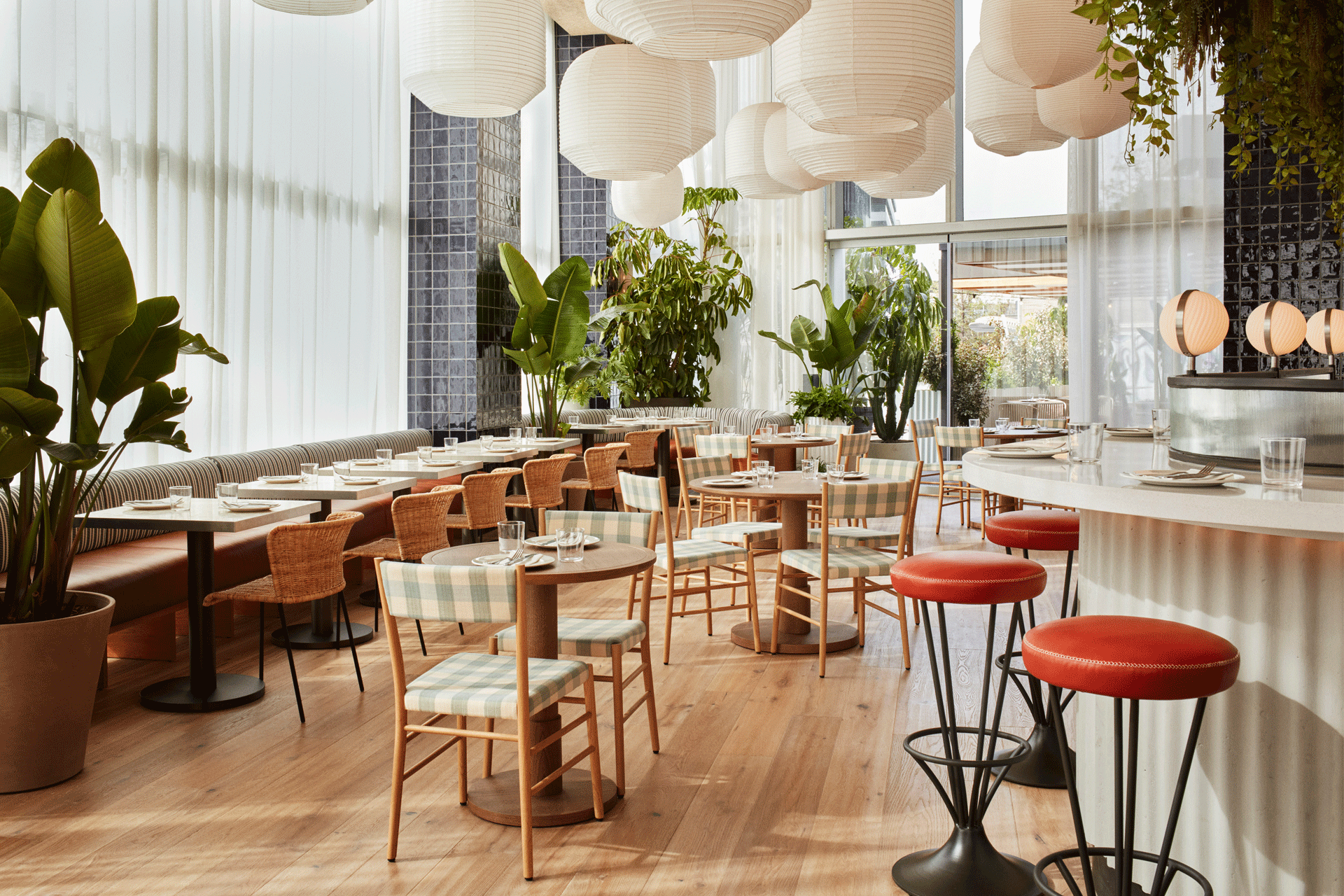 The Hoxton Poblenou, Barcelona, Spain — Light-filled restaurant with woven pendant lamps, wooden tables, rattan chairs, potted plants, and red bar stools overlooking city views.