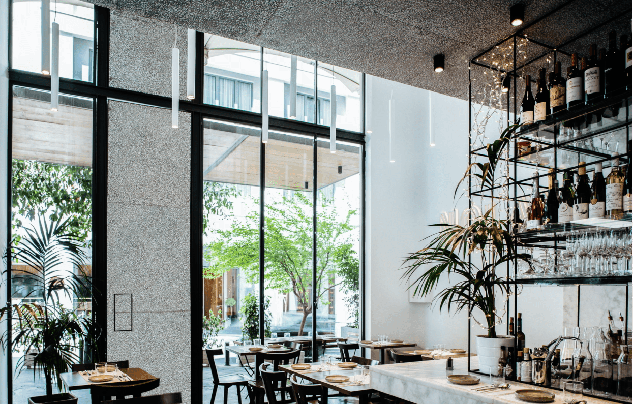 Nolan, Europe — Modern restaurant with floor-to-ceiling windows, terrazzo walls, marble bar, black metal shelving, natural light, minimalist dining setup.