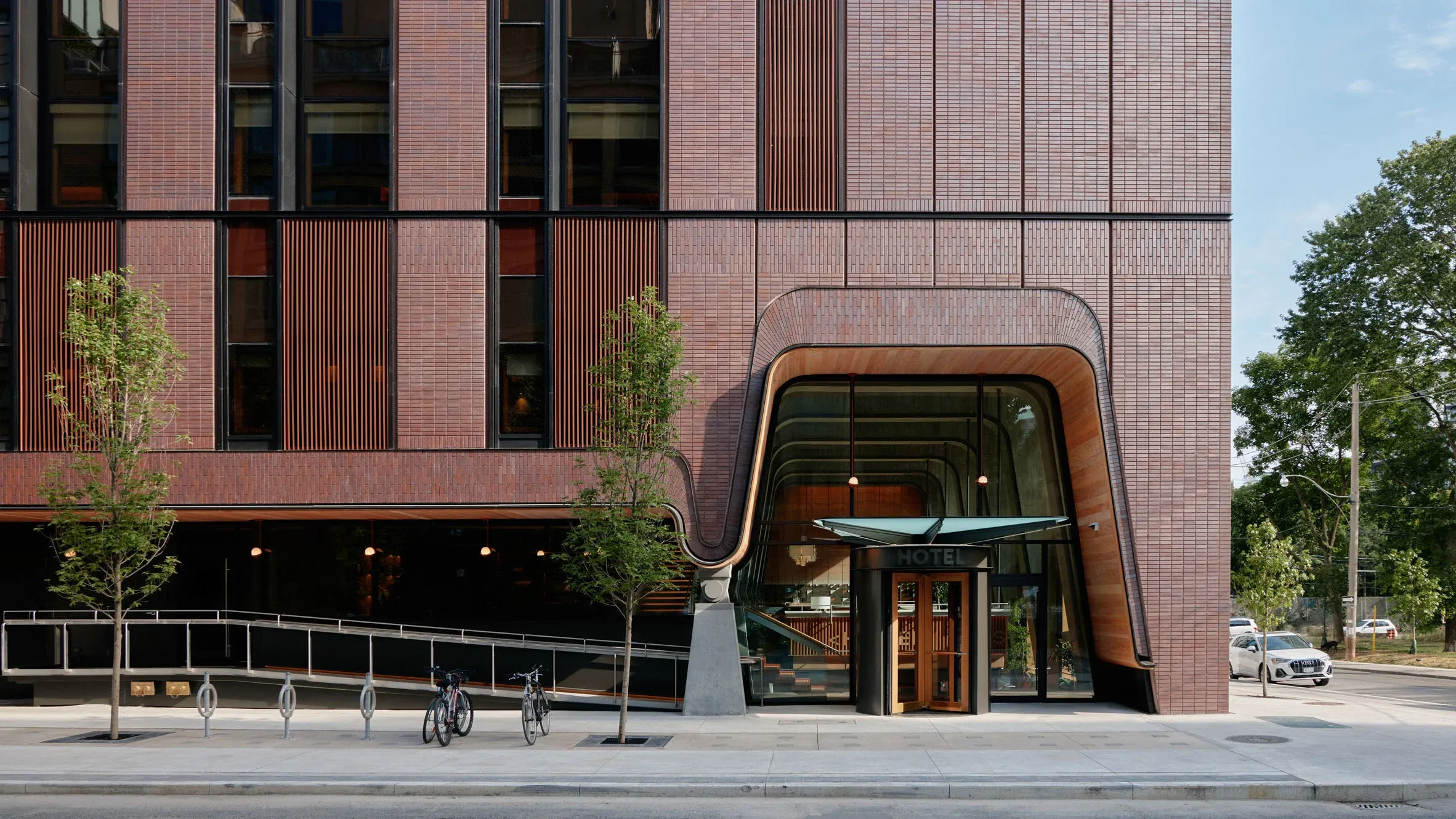 Ace Hotel Toronto, Canada — Curved copper-clad entrance with floor-to-ceiling windows, red brick facade, and street-level bicycles under afternoon light.