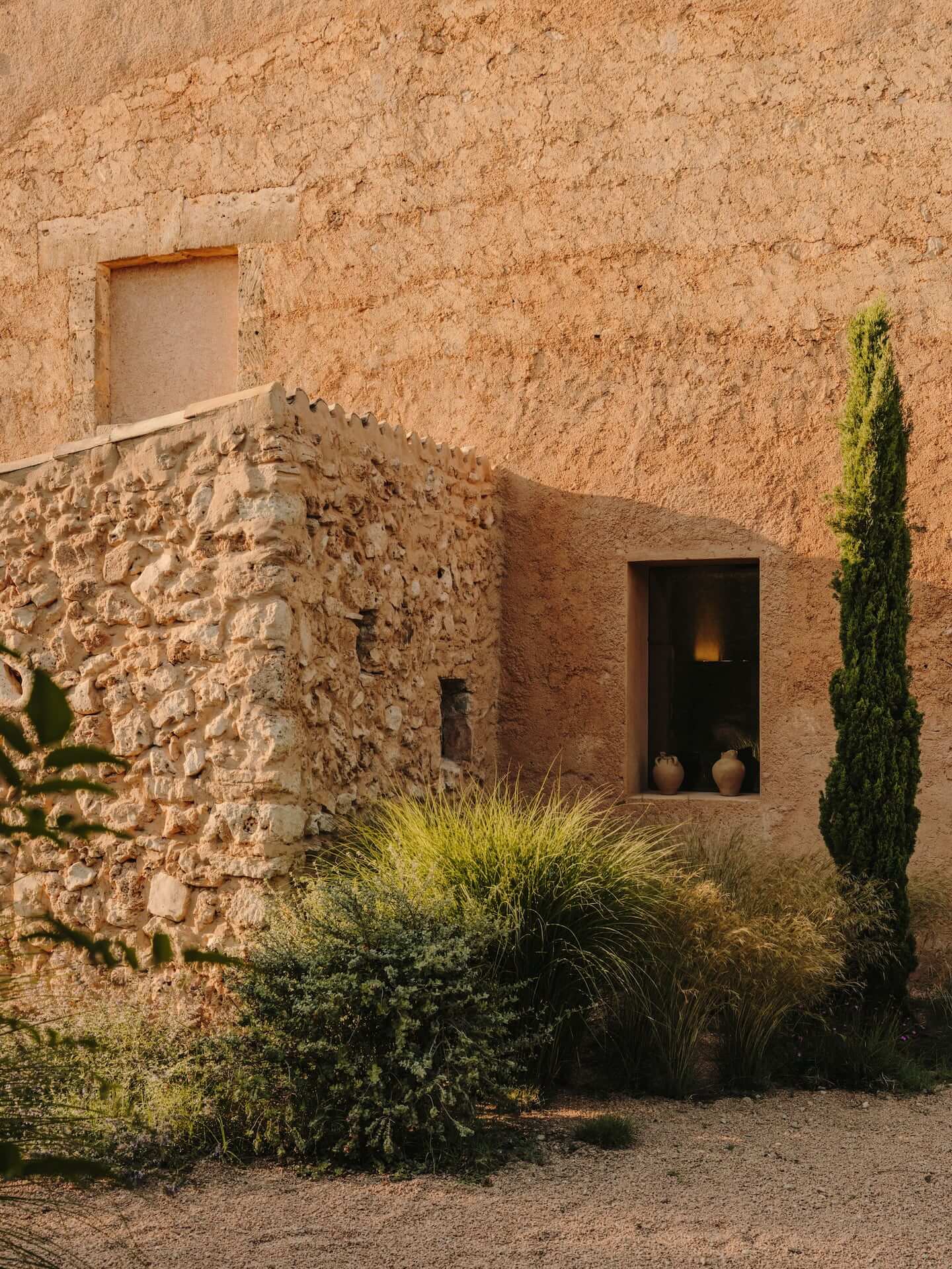 Can Lluïssó, Balearic Islands, Spain — Terracotta stone villa facade with narrow window, ceramic vessels, cypress and ornamental grasses, Mediterranean courtyard.