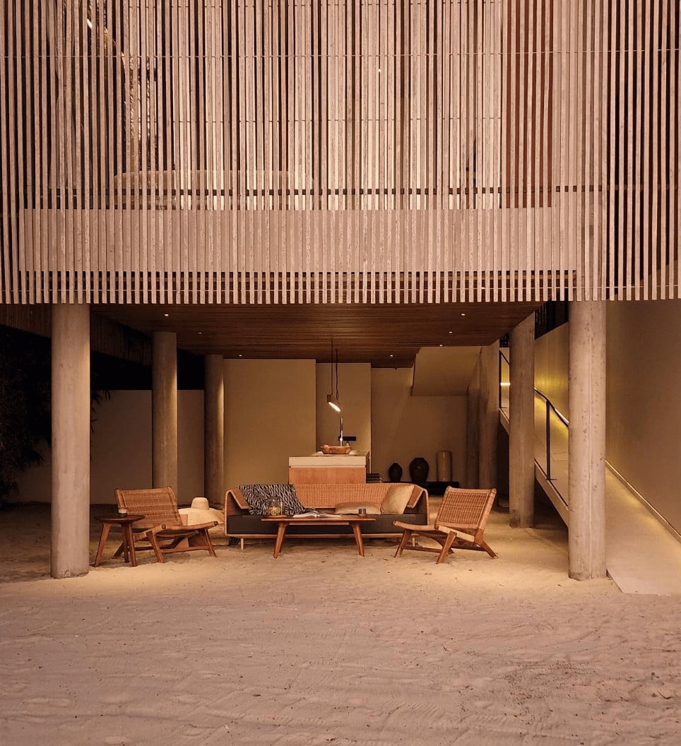 Innit Lombok, Asia — Concrete pavilion with vertical timber screens, sand floor, mid-century modern furniture, and warm ambient lighting at dusk.