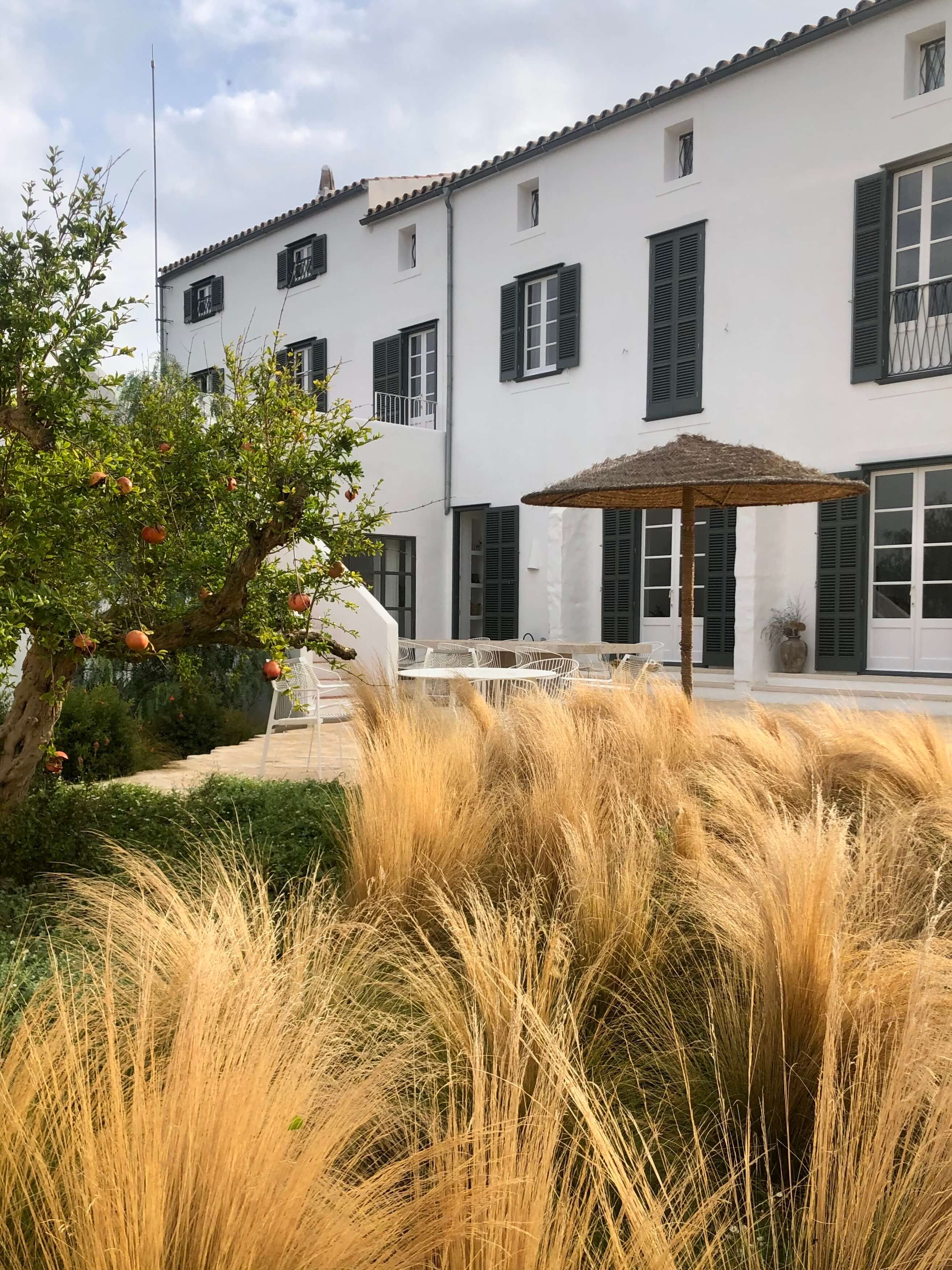 Es Bec d’Aguila — Es Bec d'Aguila, Balearic Islands, Spain — White Mediterranean villa with dark shutters, golden ornamental grasses, pomegranate tree, and thatched umbrella on paved terrace.