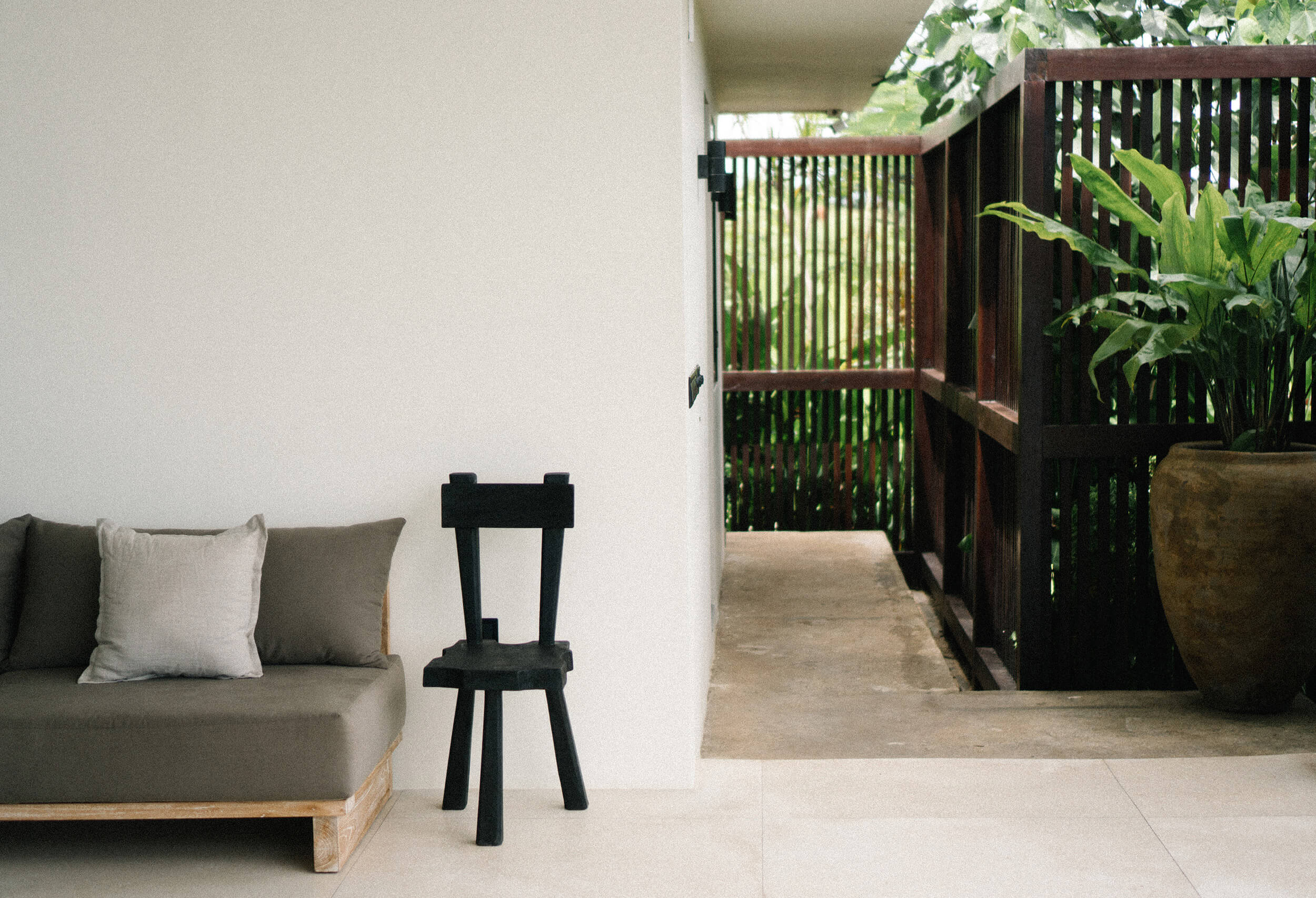 Somewhere Lombok, Asia — Minimalist covered terrace with natural linen sofa, black wooden chair, polished concrete floors, and views to lush tropical garden.