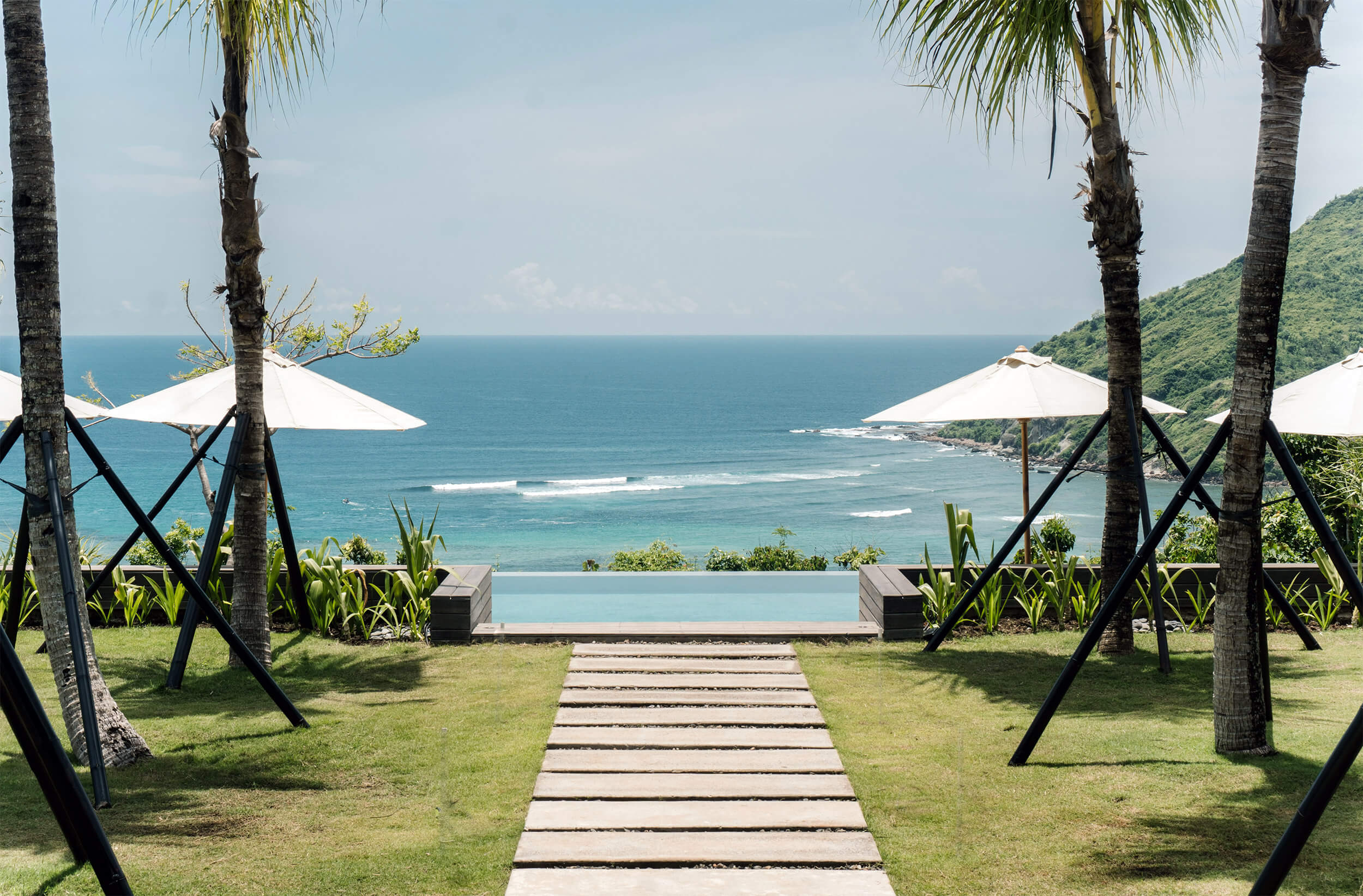 Somewhere Lombok, Lombok, Indonesia — Oceanfront infinity pool with palm trees, white umbrellas, and terraced wooden walkway overlooking turquoise waters and coastal cliffs.