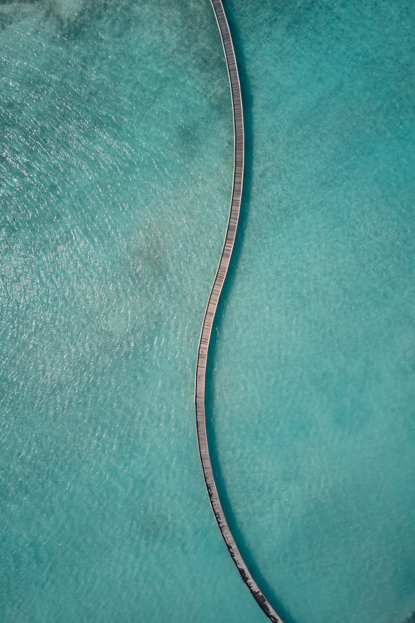 Dive Butler, Asia — Aerial view of wooden jetty extending into crystalline turquoise water, minimalist luxury resort architecture and serene tropical destination aesthetic.