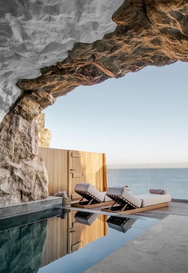 ACRO Suites, Europe — Coastal cave suite with wooden loungers, infinity pool, and sea views beneath natural stone overhang at sunset.