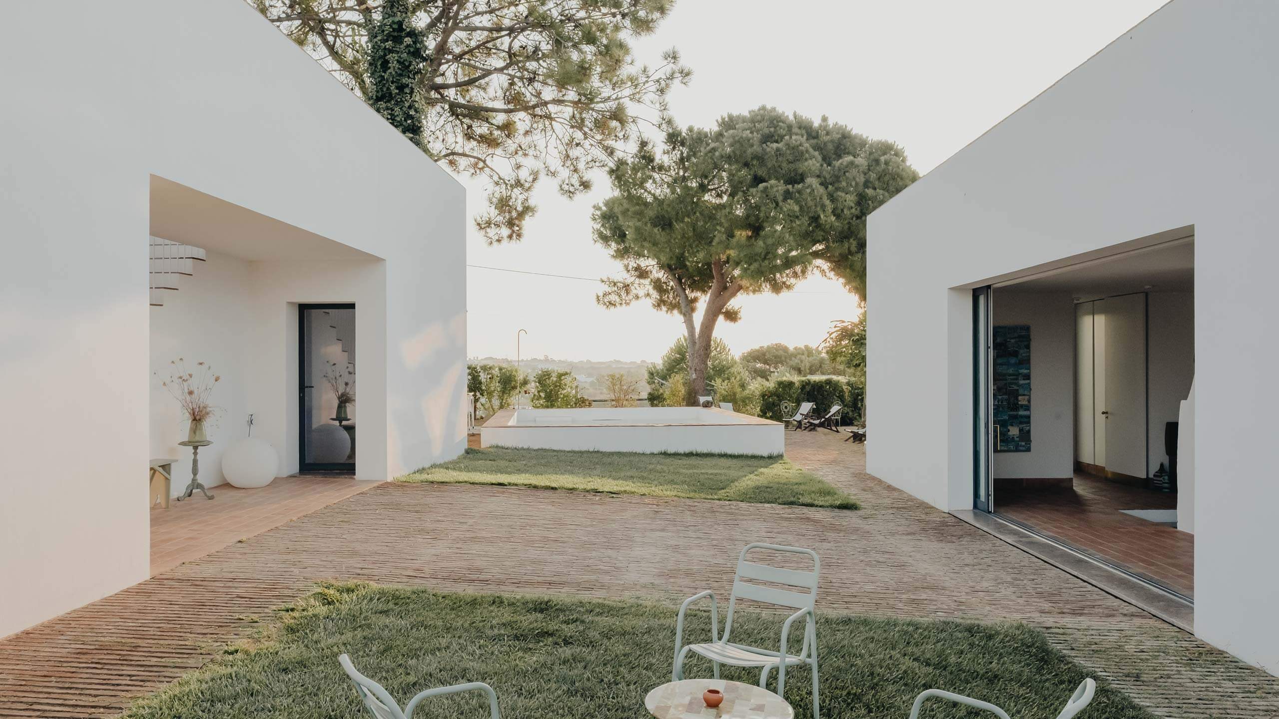 Casa Modesta, Europe — Minimalist courtyard with white cubic architecture, wooden deck, mature Mediterranean tree, and distant hillside views at golden hour.