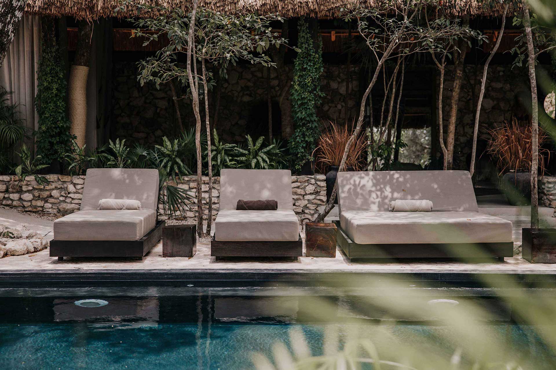 Bardo, Mexico — Three stone loungers with linen cushions beside dark infinity pool, framed by tall trees and ivy-covered walls under thatched roof.