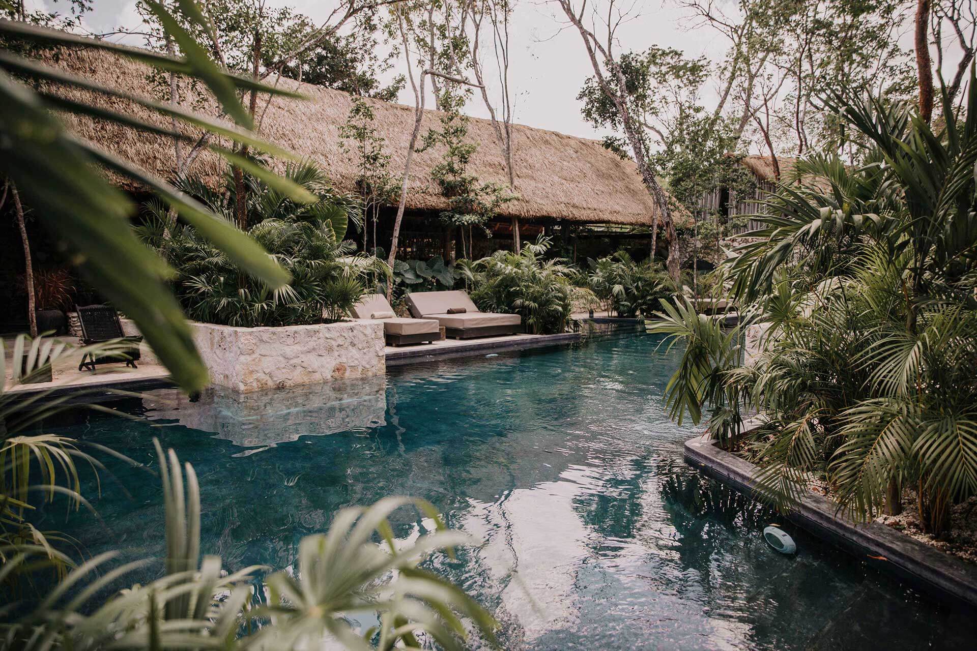 Bardo, Mexico — Tropical pool with thatched-roof pavilions, limestone terrace, palm fronds, and lounge chairs nestled in lush jungle setting.
