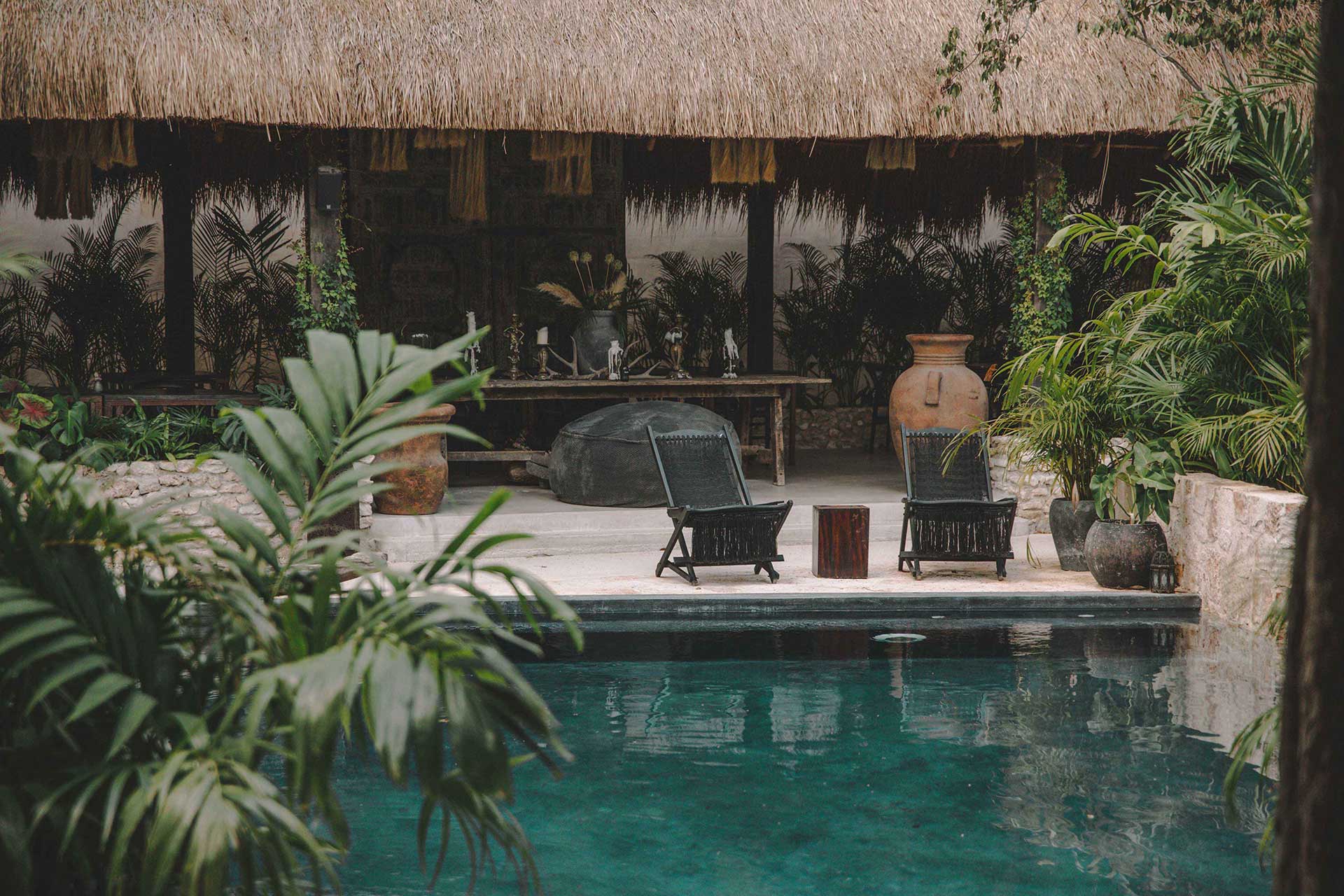 A poolside terrace at Bardo with thatched roof pavilion, dark wood loungers, and lush tropical plantings framing turquoise water.