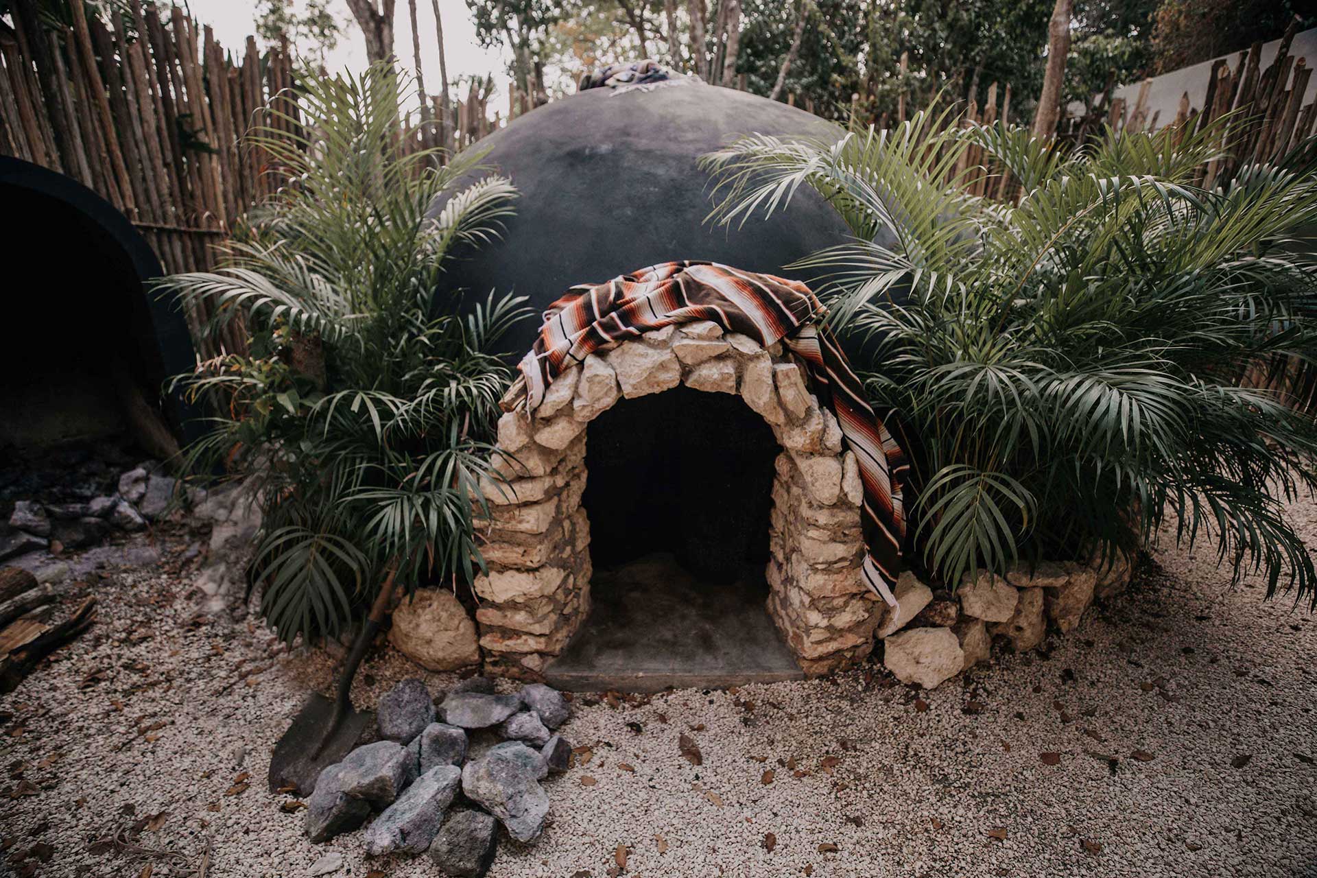 Bardo, Mexico — Stone archway with decorative red tile trim framed by palm fronds, desert sand ground, and thatched wooden structures beyond.