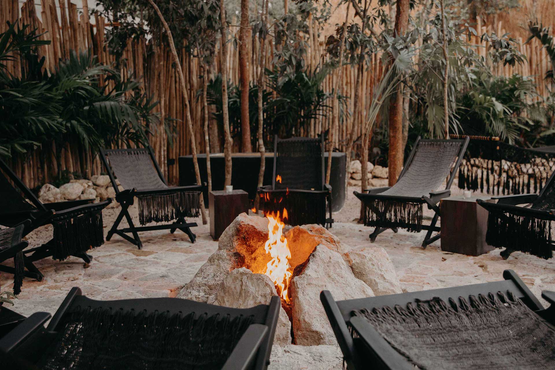 Bardo, Mexico — Firelit sandy courtyard with black lounge chairs, palm-thatched pavilions, and glowing stone fire pit at dusk.