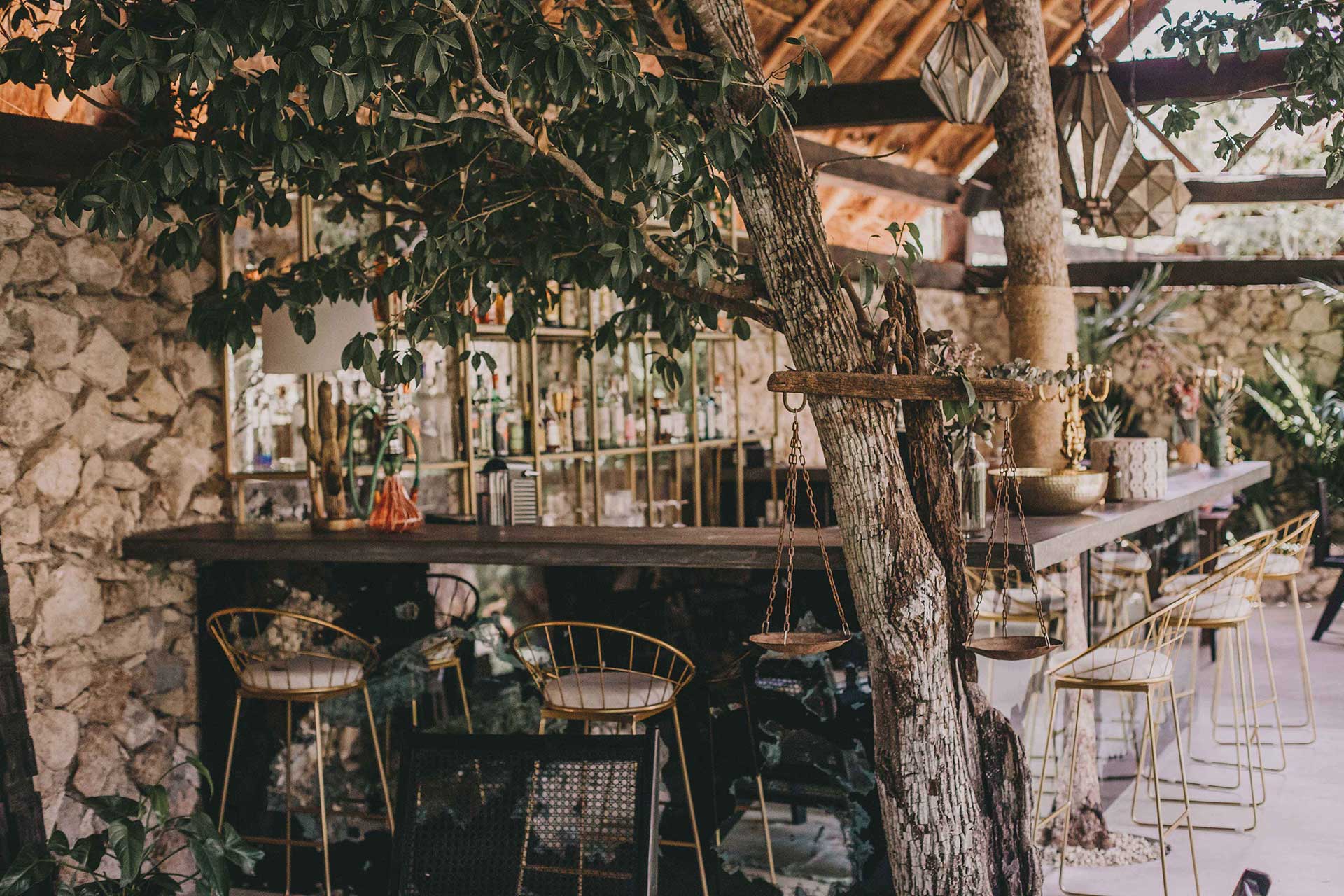 Bardo, Mexico — Wooden pergola shades stone courtyard bar with brass pendant scales, woven chairs, and mature tree creating dappled light and layered architectural depth.