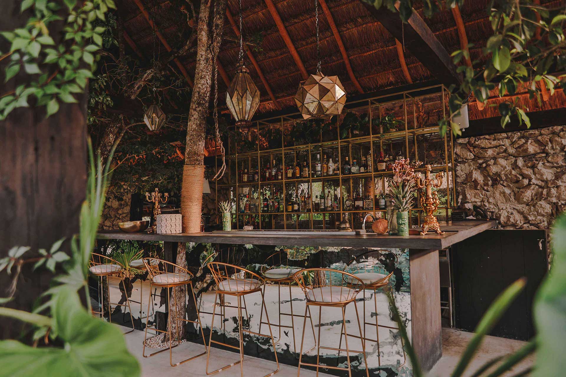 Bardo, Mexico — Thatched-roof bar pavilion with geometric pendant lights, brass bar stools, stone walls, and lush tropical vegetation creating intimate evening ambiance.