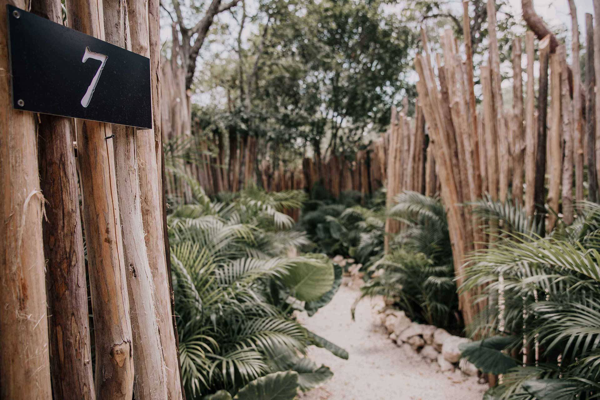Bardo, Mexico — Wooden pergola with number 7 marker overlooking palm-lined pathway through lush tropical garden with tall timber posts and green vegetation.