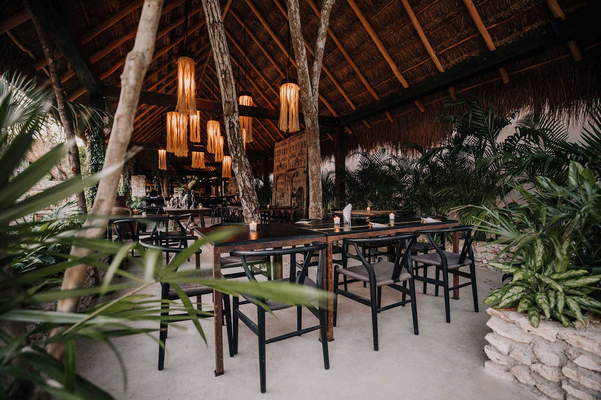 A thatched-roof dining pavilion at Bardo features wooden tables, black chairs, and woven pendant lamps among towering palms and natural timber posts.