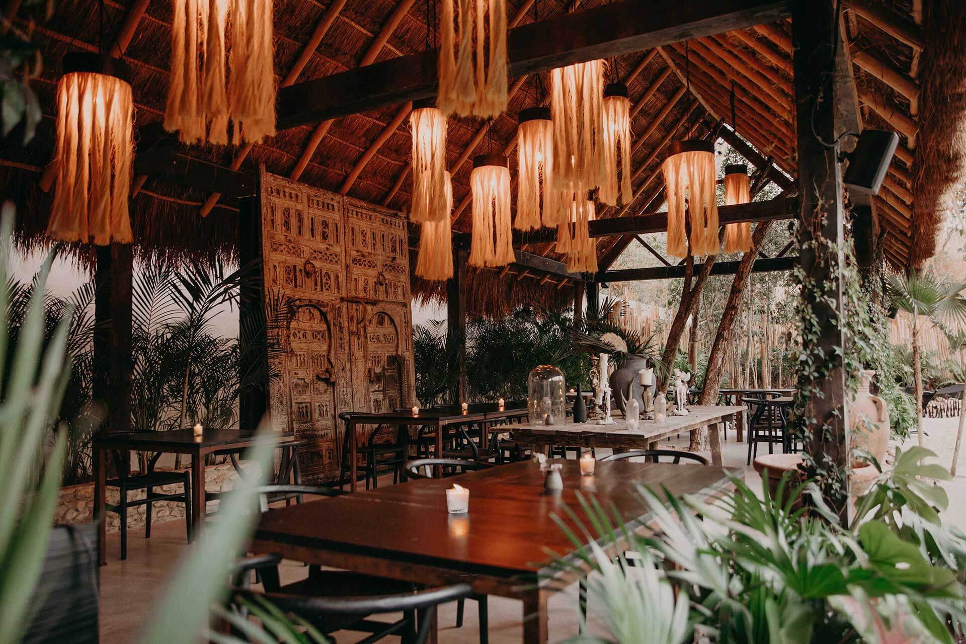 Bardo, Mexico — Thatched-roof dining pavilion with carved wooden panels, pendant lighting, tropical plants, and candlelit tables in warm amber tones.