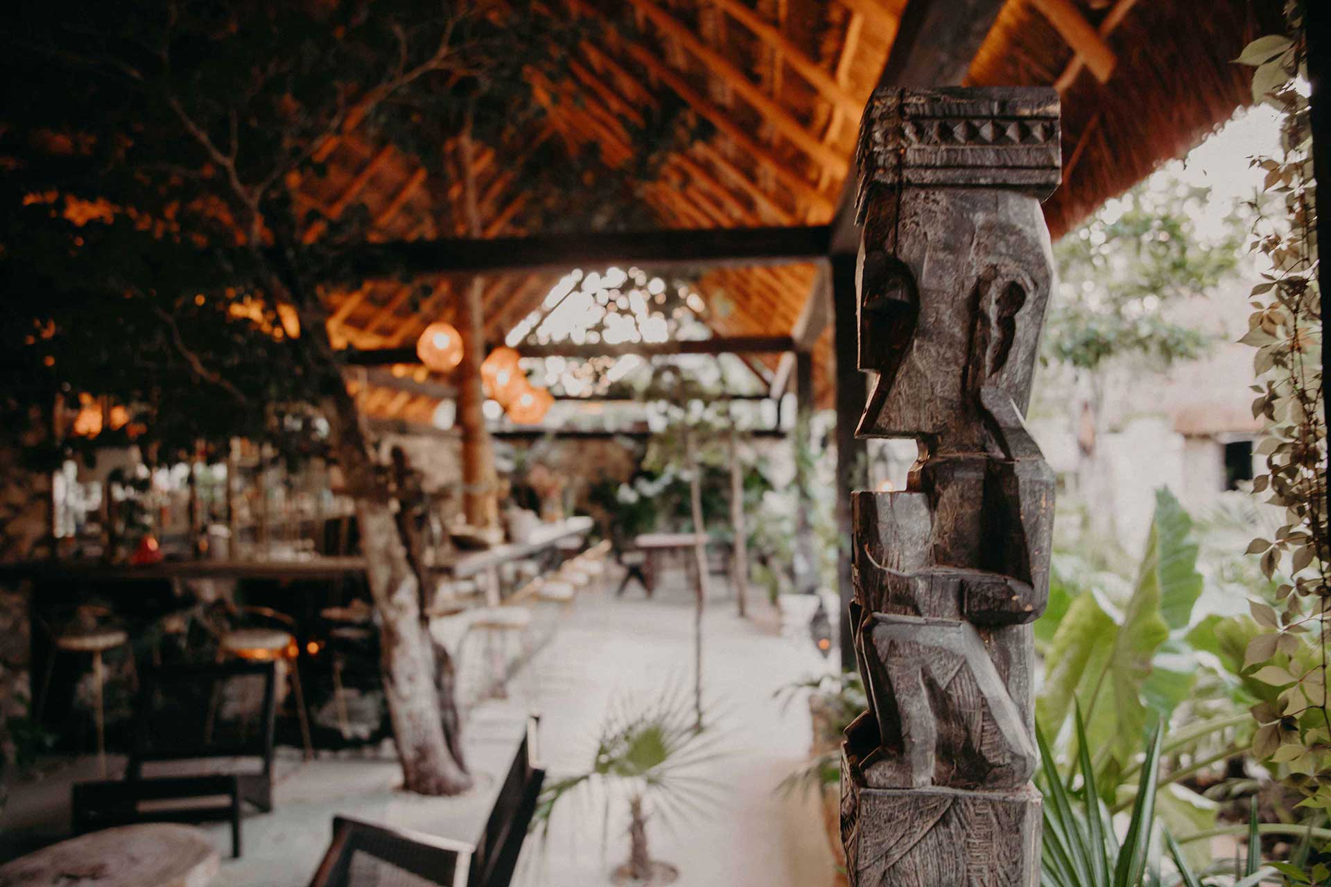 Bardo, Mexico — Carved wooden column framing open-air dining pavilion with warm string lights, stone courtyard, and lush vegetation at dusk.