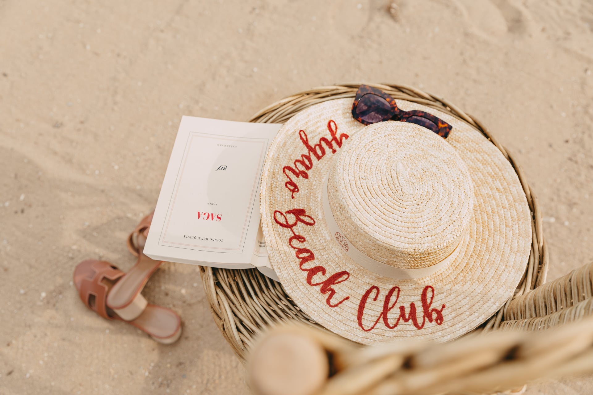 Lily of the Valley, Europe — Woven straw hat with red embroidered script beside white luxury brochure and tortoiseshell sunglasses on sand.