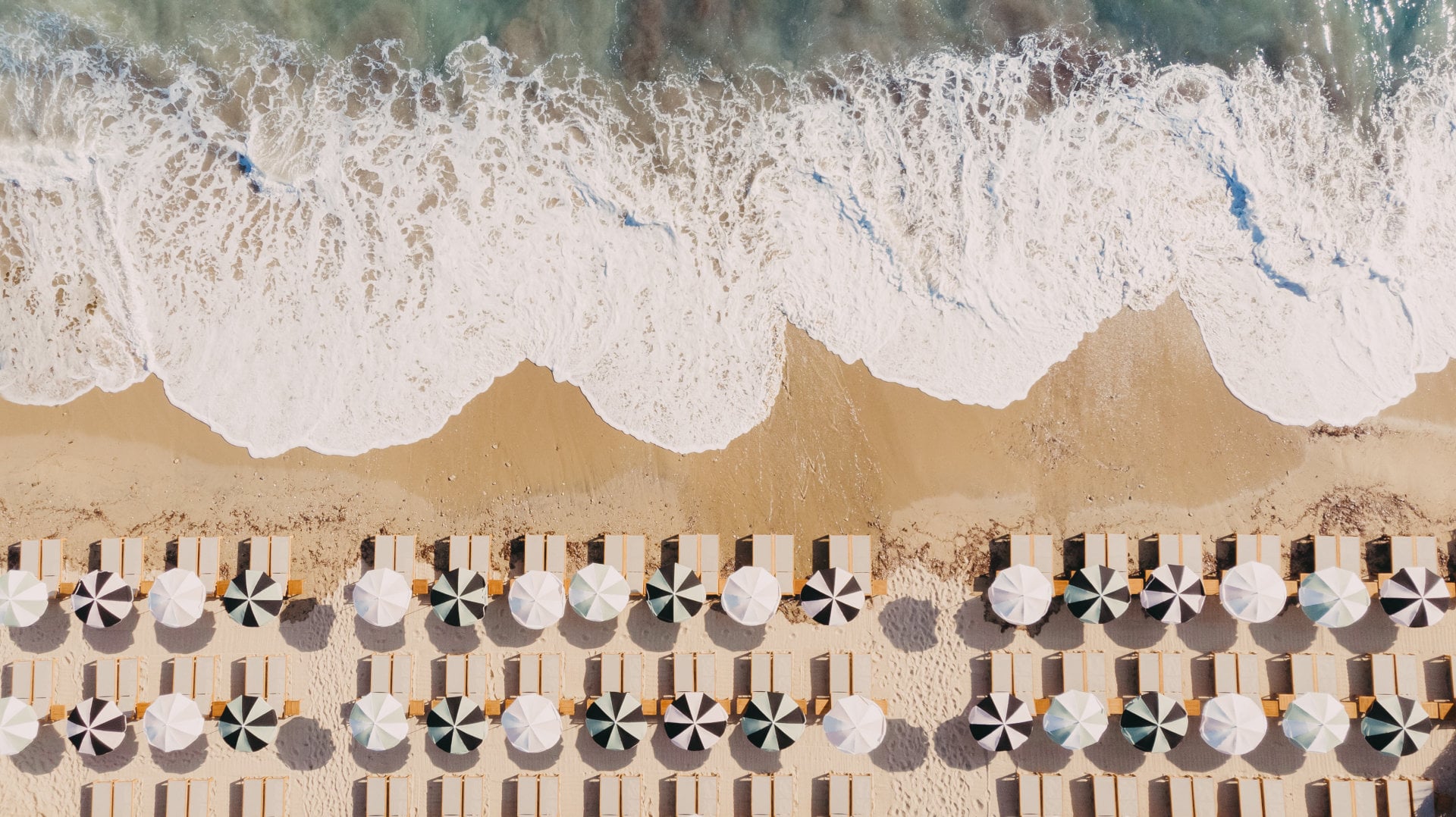 Lily of the Valley, Europe — Overhead view of striped beach loungers and black-and-white umbrellas arranged in rows on sand with waves breaking along the shore.
