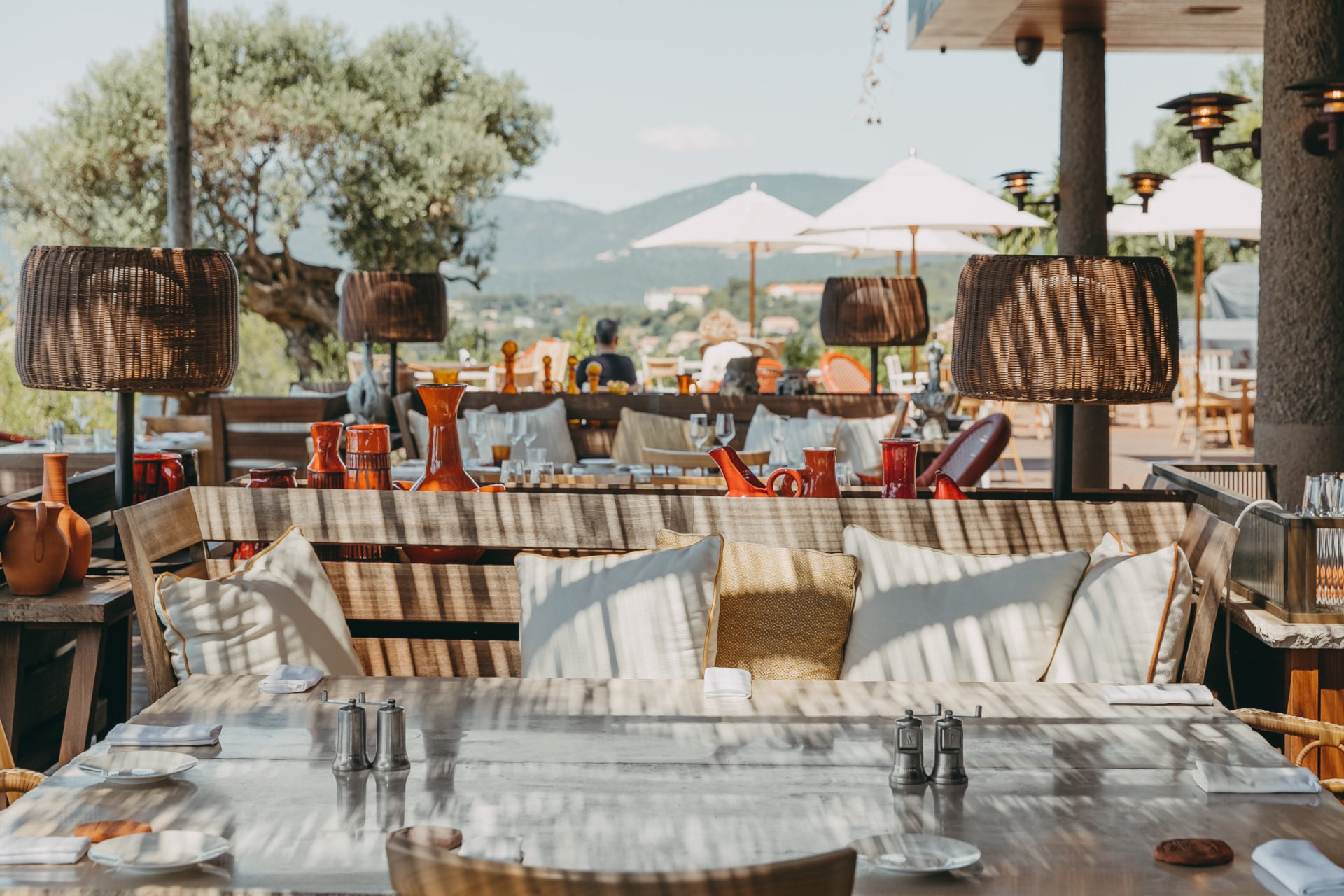 Lily of the Valley, Europe — Woven pendant lights and striped cushions frame a marble dining table overlooking Mediterranean hills and olive trees.