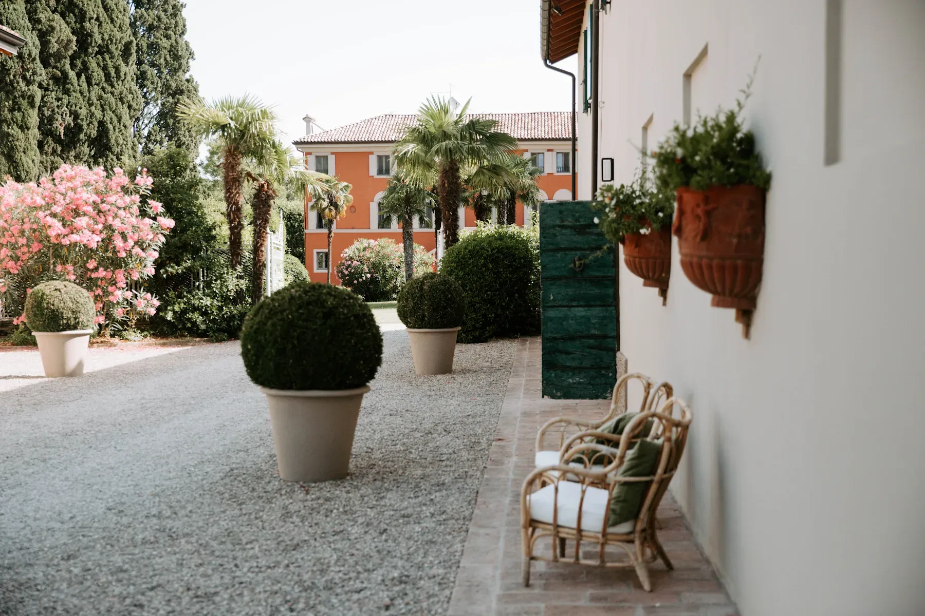 Roncolo 1888, Europe — Courtyard with boxwood topiaries, pink flowering shrubs, palm trees, ochre villa, gravel terrace, wicker chairs, Mediterranean garden design.