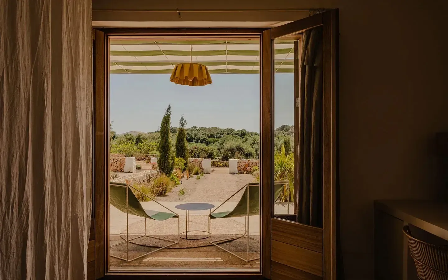 Amagatay, Balearic Islands — Open terrace with olive-green hammock chairs, golden pendant lamp, striped awning, Mediterranean landscape views, warm stone and wood interior.