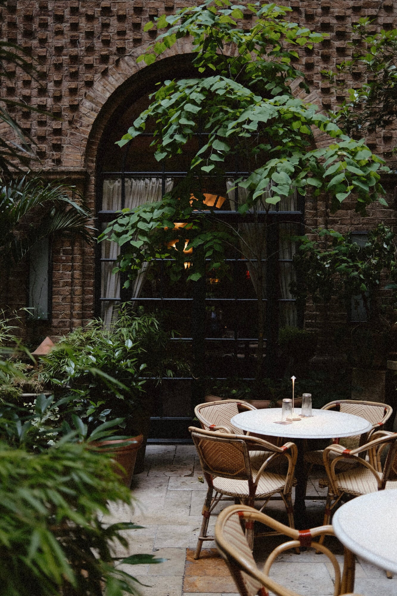 The Greenwich Hotel, New York, United States — Brick courtyard with ivy-draped arches, rattan bistro chairs, marble tables, and warm interior lighting at dusk.