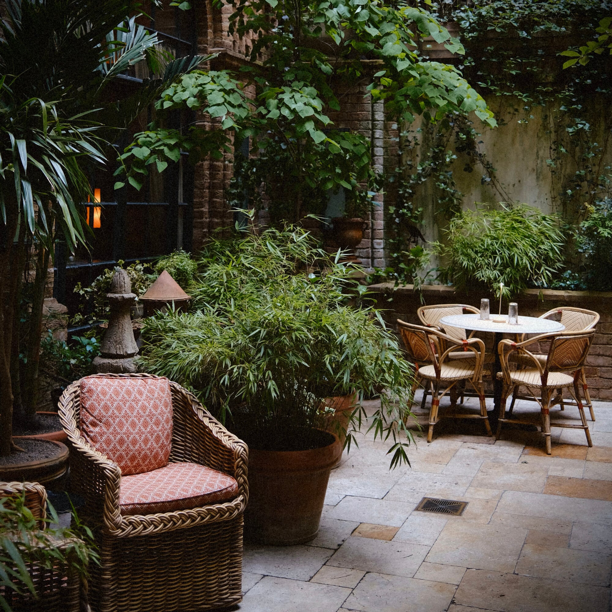 The Greenwich Hotel, New York, United States — Ivy-draped courtyard with woven rattan chairs, stone paving, potted bamboo, and brick facade evoking classic European garden aesthetic.