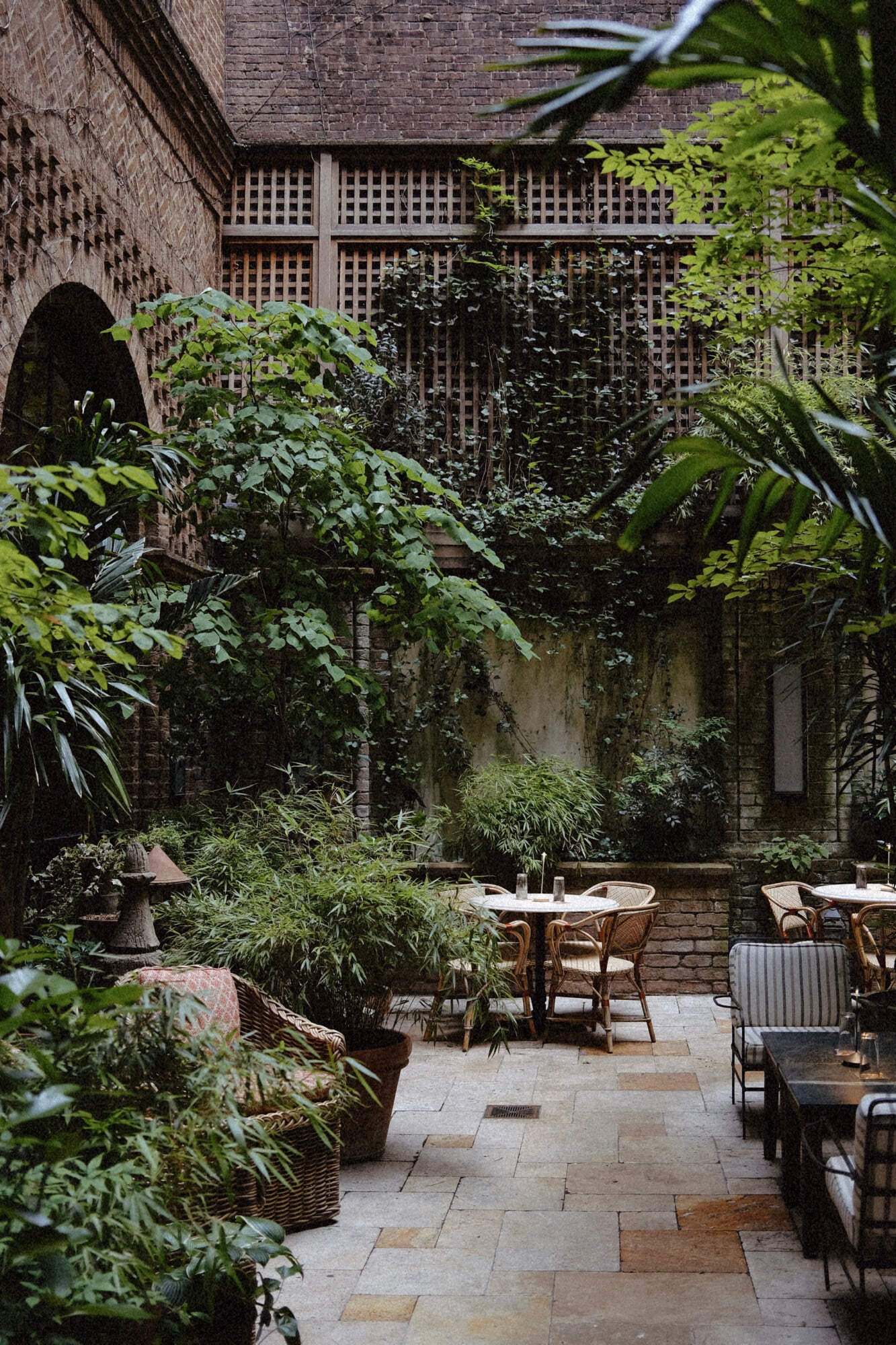 The Greenwich Hotel, New York, USA — Ivy-draped courtyard with brick walls, lattice trellis, bistro seating, and lush potted plants in dappled daylight.
