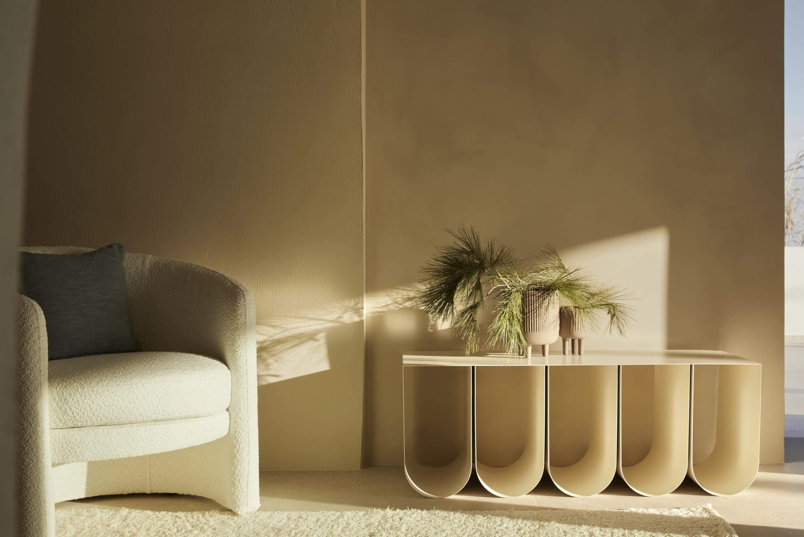 Pārocks, Europe — Minimalist hospitality interior with curved cream armchair, sculptural console table, potted palm, and warm natural light casting geometric shadows on ochre walls.