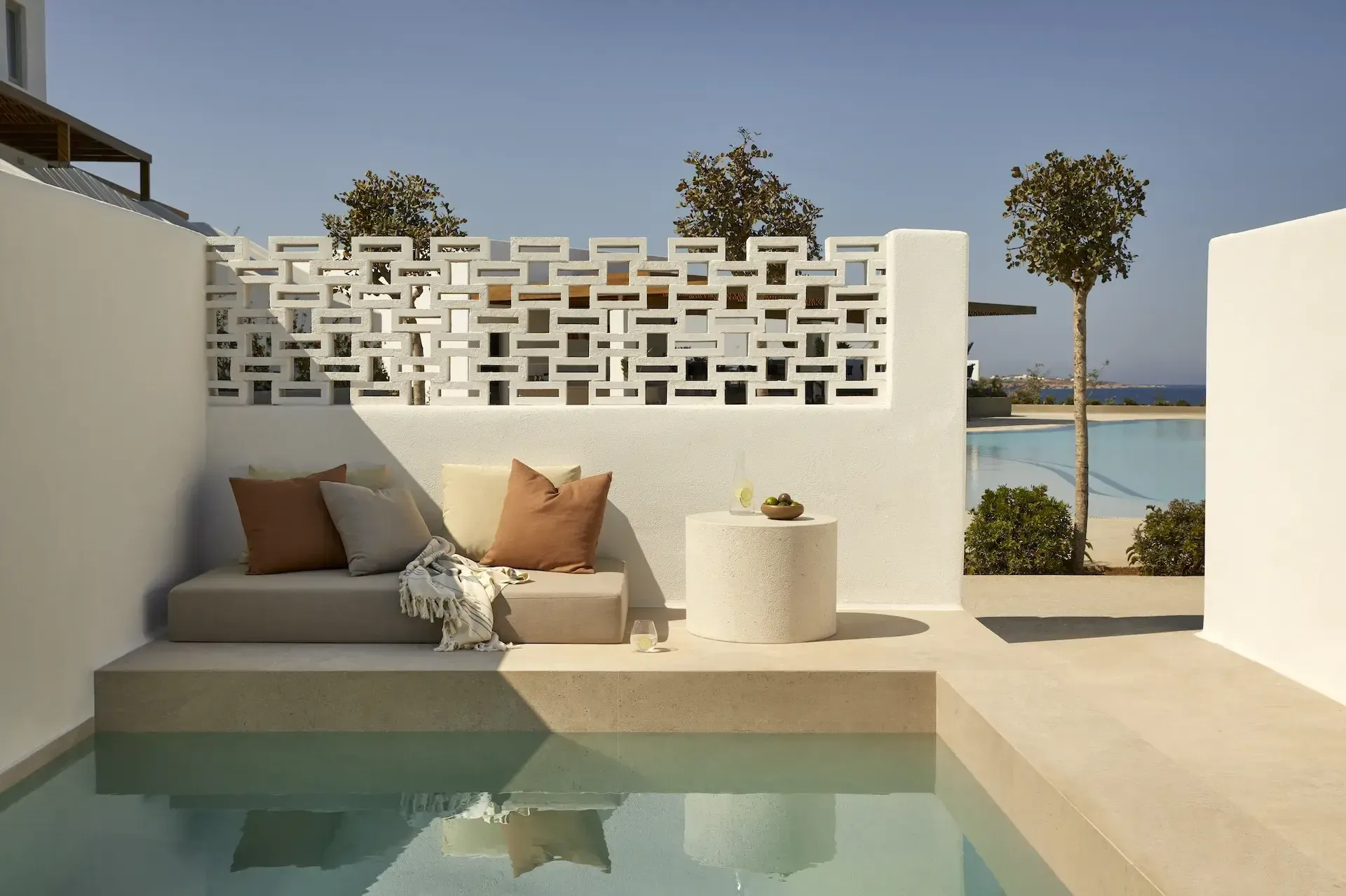 Pārocks, Europe — Minimalist white poolside terrace with geometric screen wall, sunlit seating nook, calm water reflection, and Mediterranean coastal view.