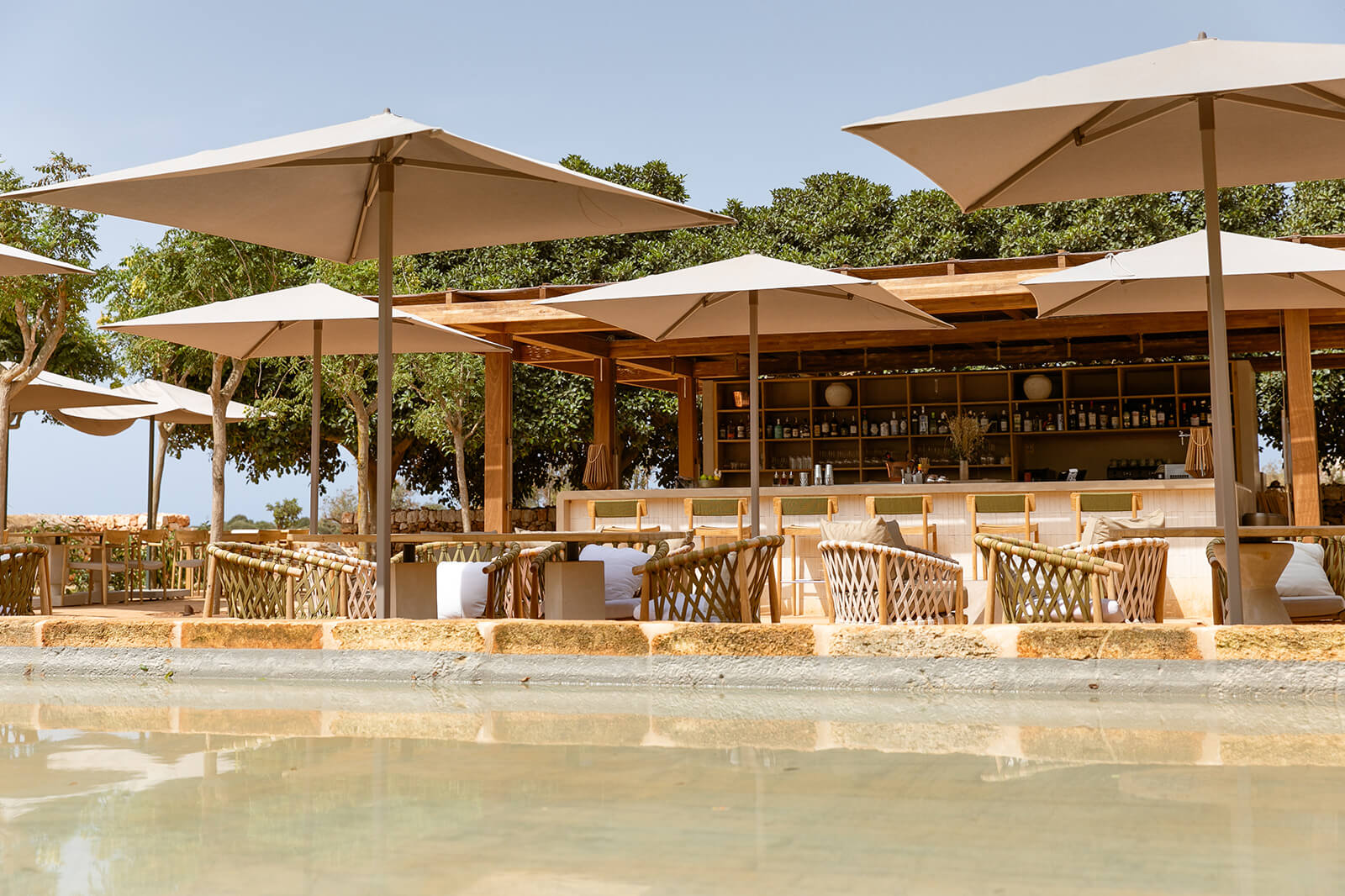 Sa Clarisa, Balearic Islands — Beachfront restaurant pavilion with timber framework, canvas umbrellas, woven chairs, and open-air bar overlooking shallow turquoise water.