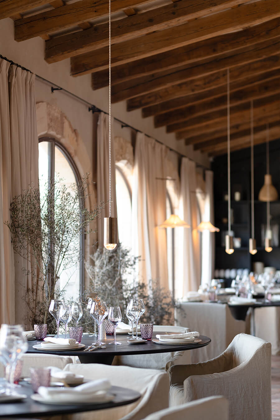 Vermell, Balearic Islands — Dining room with exposed wooden beams, pendant lights, linen curtains, round black table, and minimalist table settings overlooking Mediterranean light.