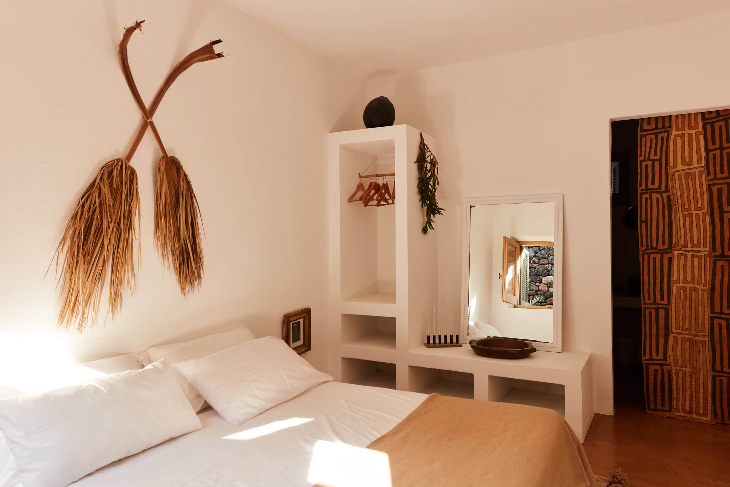 Parco Dei Sesi, Europe — Minimalist bedroom with white walls, wooden accents, dried palm decoration, and geometric patterned door in warm earth tones.