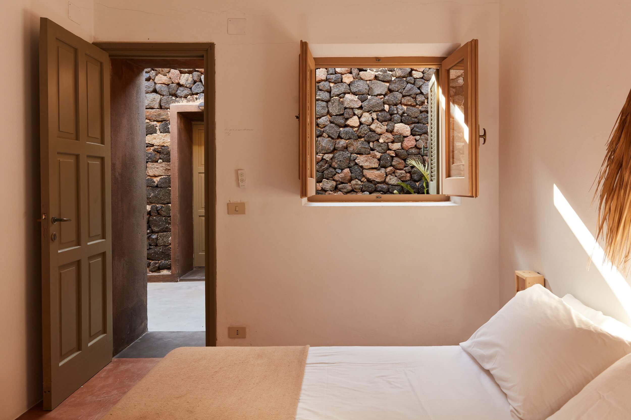 Parco Dei Sesi, Europe — Bedroom with terracotta walls, wooden doors, open window framing stone wall, natural light, minimalist Mediterranean design.