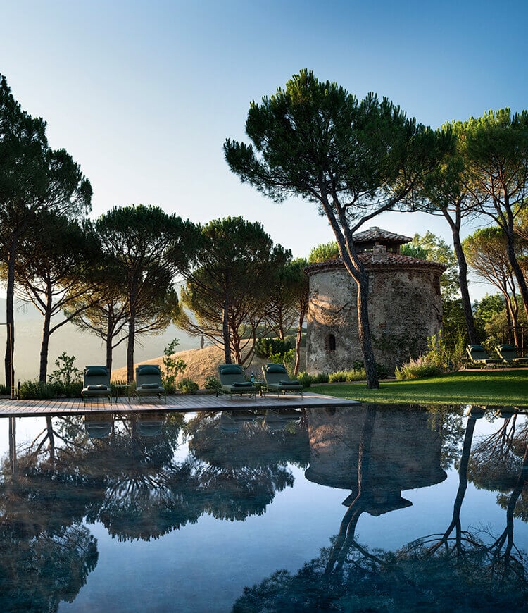 Il Torrino, Europe — Stone tower and umbrella pines reflected in still water, with lounge chairs on deck at dusk, Mediterranean landscape architecture.