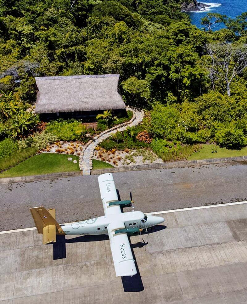 Islas Secas, Panama — Seaplane on runway beside thatched-roof bungalow nestled in tropical forest with ocean beyond.