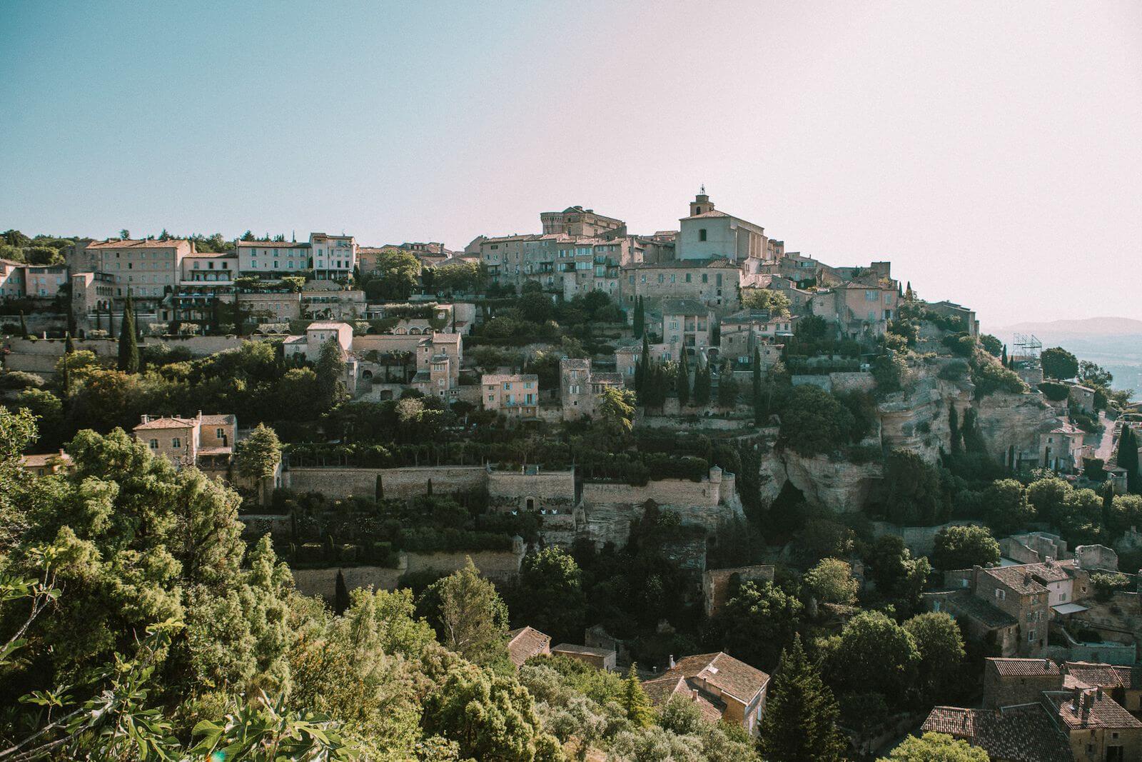 Gordes, France — Hilltop village with ochre stone buildings, cypress trees, and medieval architecture cascading down a limestone cliff at golden hour.